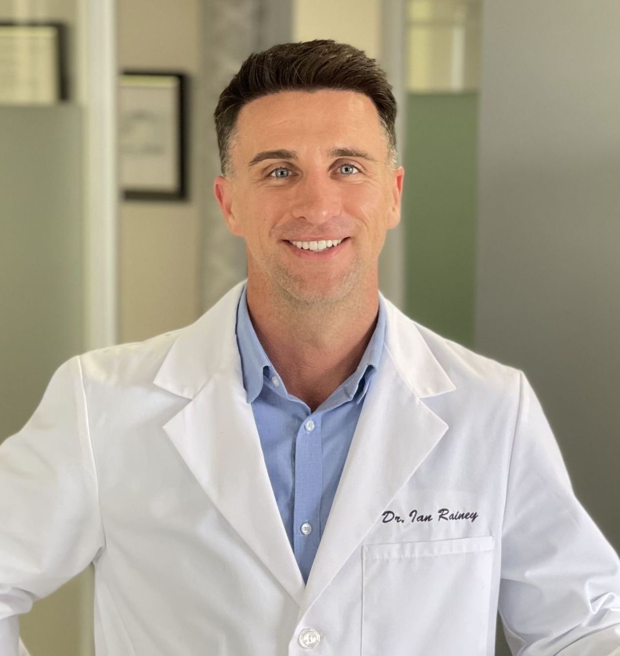 Dr. Rainey Dr. Rainey of Rainey Chiropractic, smiling and standing with left arm resting on a half wall with right hand on hip; wearing blue shirt and buttoned up medical jacket.